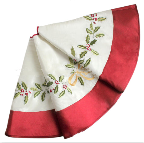 White holly embroidered Christmas tree skirt in white on a wooden floor with presents underneath a decorated tree